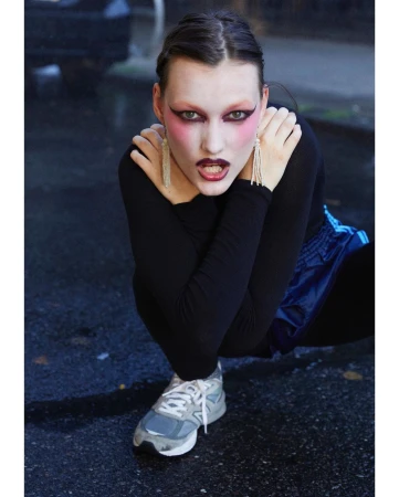 Young woman crouches on a wet urban street in black top, blue shorts, gray sneakers, pink eye shadow, and long earrings.