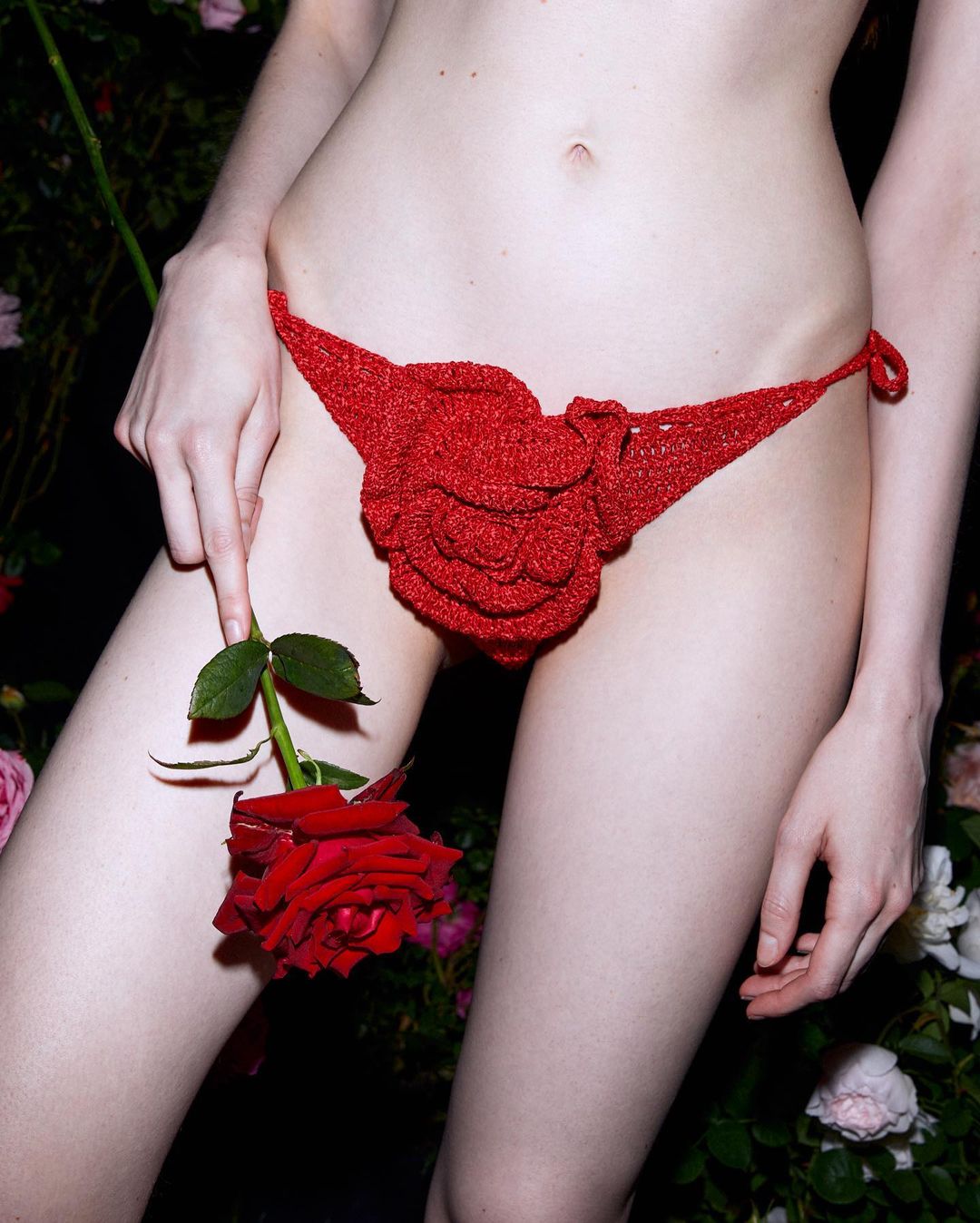 Close-up of a fair-skinned adult female wearing a red crochet bikini bottom and holding a red rose against a floral background.