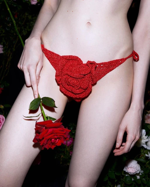 Close-up of a fair-skinned adult female wearing a red crochet bikini bottom and holding a red rose against a floral background.