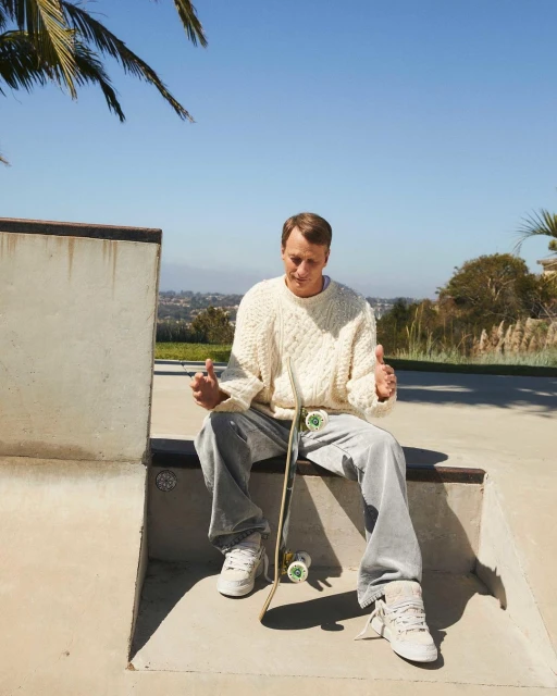 Man sits with skateboard in skatepark under blue sky. He wears cream sweater, gray pants, and white sneakers in daytime setting.
