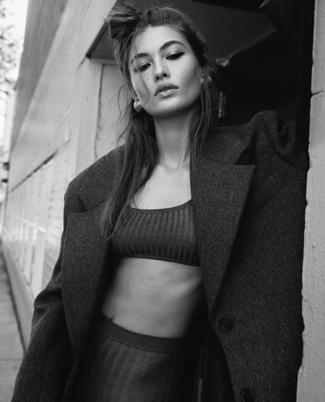 Black and white portrait of a woman wearing an oversized wool coat and crop top, leaning against an exterior wall in New York