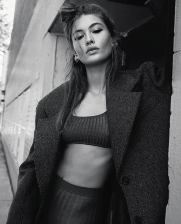 Black and white portrait of a woman wearing an oversized wool coat and crop top, leaning against an exterior wall in New York