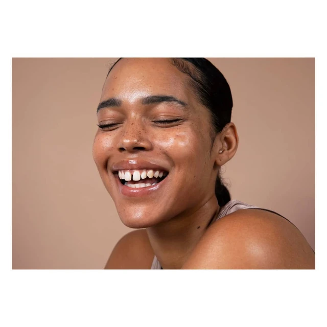 Close-up portrait of a young woman with brown skin and freckles, eyes closed, smiling broadly in front of a neutral beige background.