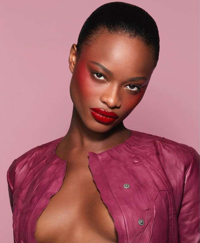 Close-up portrait of a striking dark-skinned woman with vibrant red lipstick and artistic blush, wearing a textured magenta leather jacket.
