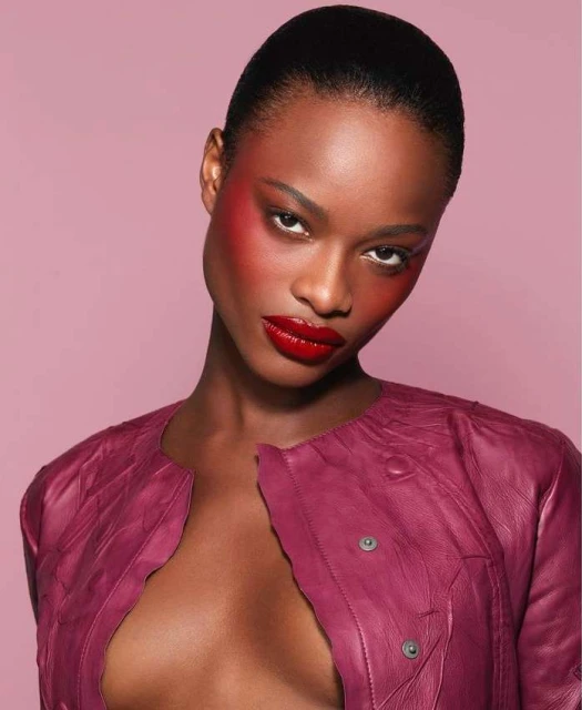 Close-up portrait of a striking dark-skinned woman with vibrant red lipstick and artistic blush, wearing a textured magenta leather jacket.