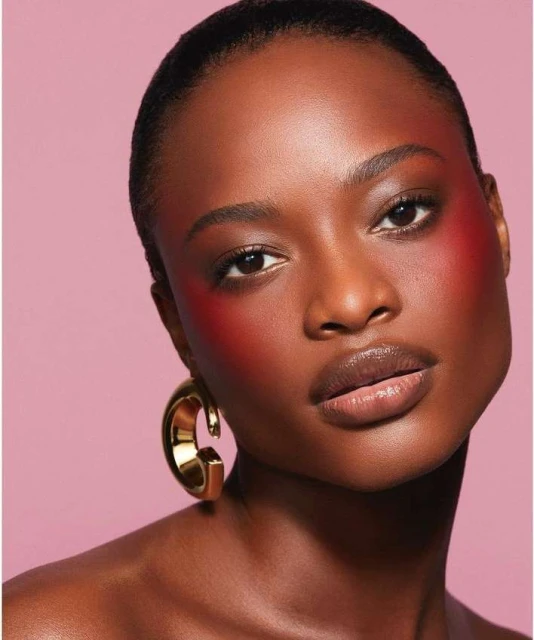 Close-up of a dark-skinned woman with red blush on her cheeks and glossy lips, wearing large gold hoop earrings against pink background.