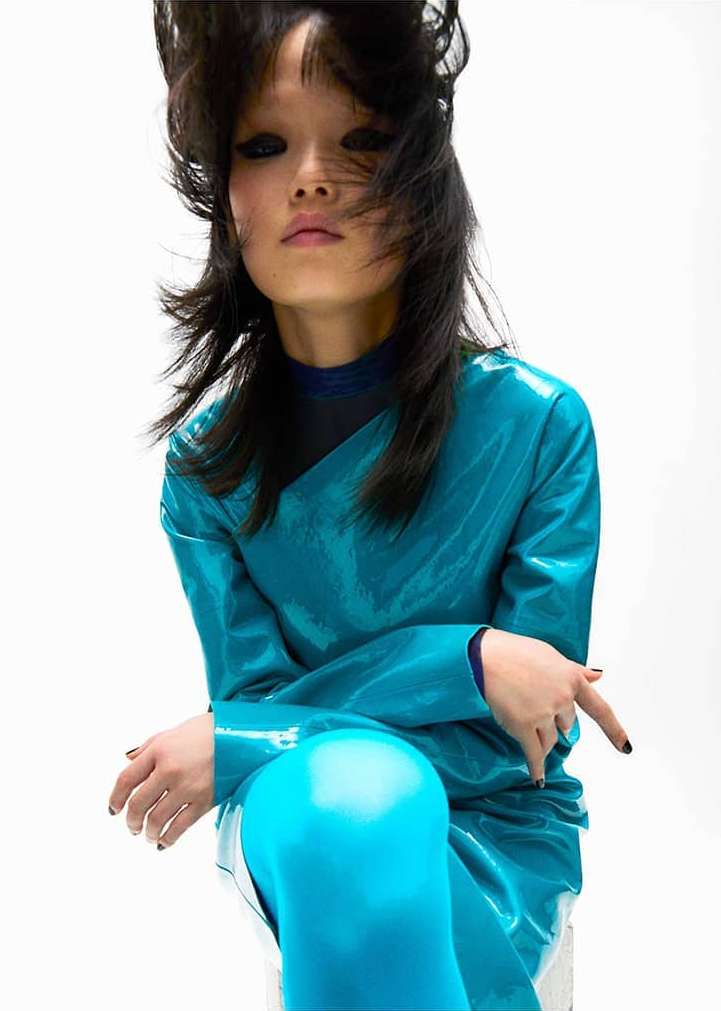 Asian woman with dark hair and glossy turquoise dress strikes a pose against a stark white backdrop in a high-fashion editorial shot.