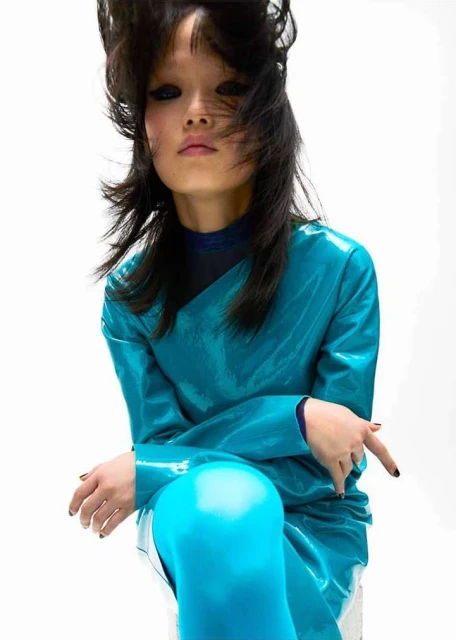 Asian woman with dark hair and glossy turquoise dress strikes a pose against a stark white backdrop in a high-fashion editorial shot.