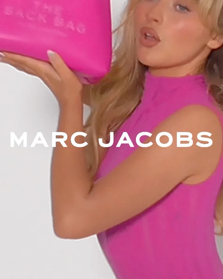 Blonde woman poses in a pink bodysuit while holding a pink purse with the logo “The Back Bag” against a plain white background.