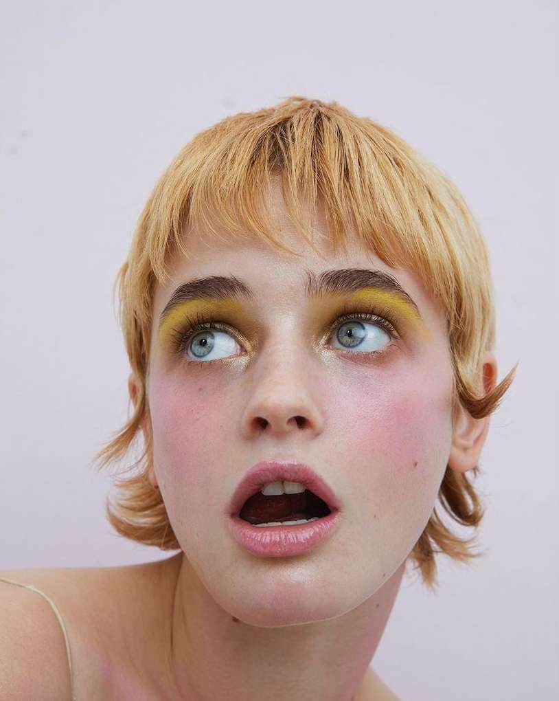 Close-up portrait of a woman with yellow eye makeup and short blonde hair looking up with her mouth slightly open against a pink background.