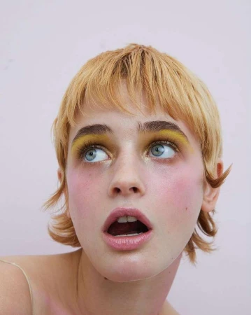Close-up portrait of a woman with yellow eye makeup and short blonde hair looking up with her mouth slightly open against a pink background.