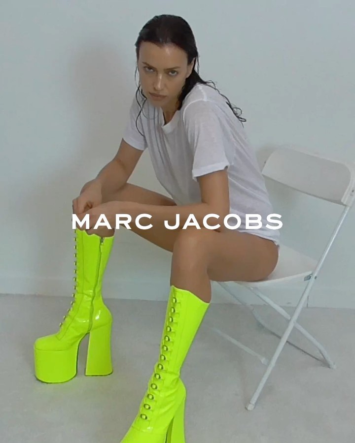 A woman with slicked-back hair poses indoors sitting on a folding chair wearing a white T-shirt and bright-green, high-platform boots.