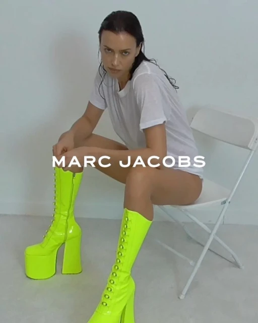 A woman with slicked-back hair poses indoors sitting on a folding chair wearing a white T-shirt and bright-green, high-platform boots.