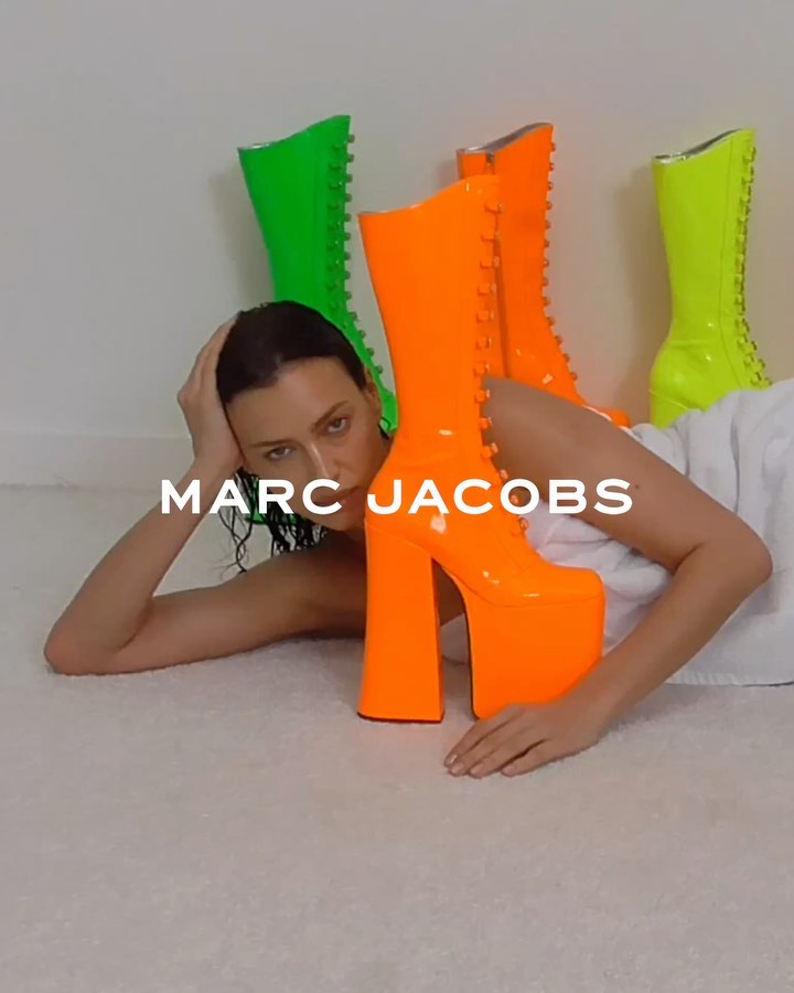 Fashion shot of a woman lying on a white surface with green and orange platform boots, Marc Jacobs logo visible, studio lighting.