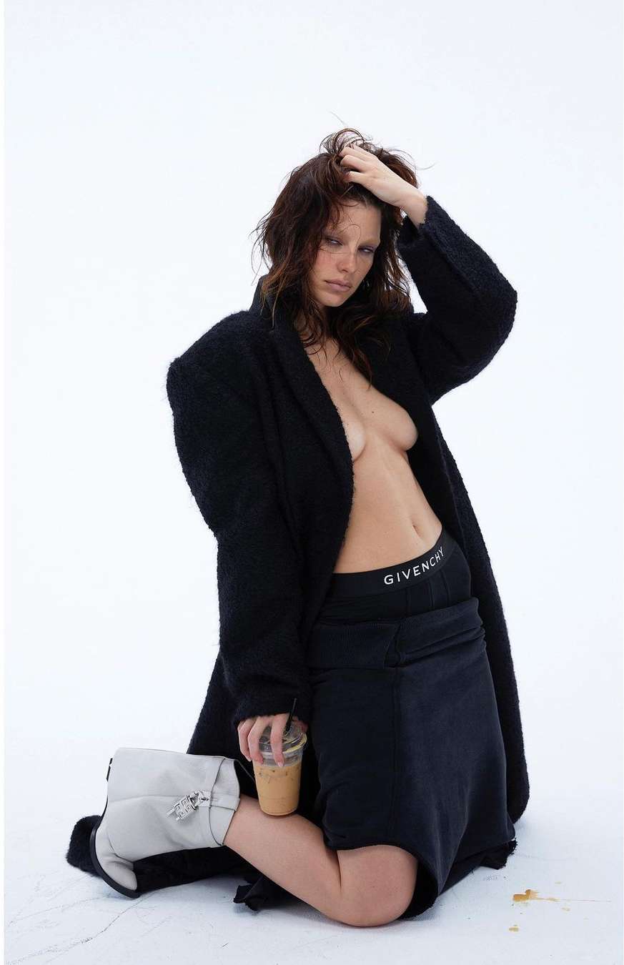 Woman in black coat poses kneeling with coffee and Givenchy underwear, set against white background in a fashion forward scene.