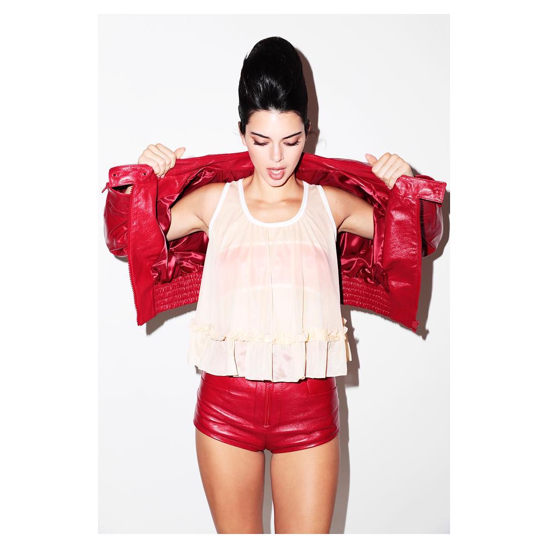 A young woman in a white top and red leather shorts poses in a photo studio, wearing a red leather jacket.