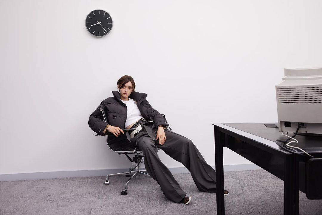A woman in a puffer jacket and grey slacks sits in an office chair in an office setting with a computer, desk, and wall clock.