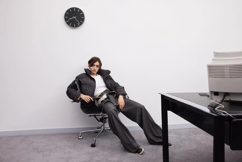 A woman in a puffer jacket and grey slacks sits in an office chair in an office setting with a computer, desk, and wall clock.