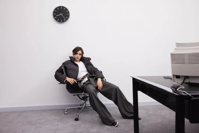 A woman in a puffer jacket and grey slacks sits in an office chair in an office setting with a computer, desk, and wall clock.