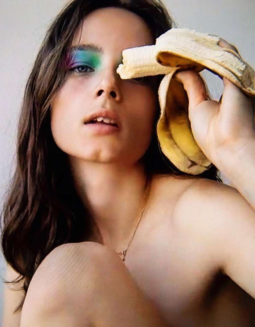 Portrait of a topless young woman with colorful makeup and dark hair holding a banana near her face against a white backdrop.