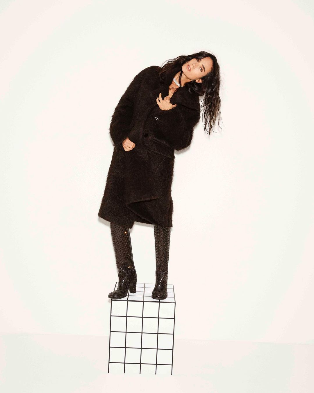 Fashionable woman posing in a long black coat and snake-print boots, leaning to her right while standing on a white grid cube.