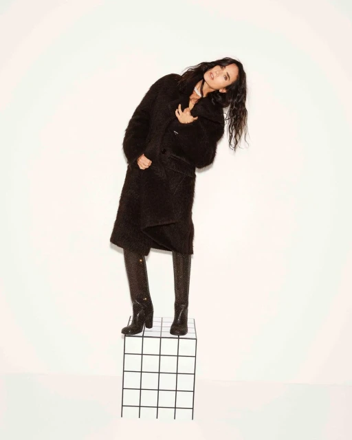 Fashionable woman posing in a long black coat and snake-print boots, leaning to her right while standing on a white grid cube.