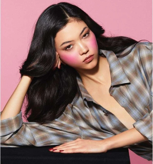 Portrait of a young Asian woman with vibrant pink blush, dark hair, and a casual plaid shirt, posing in front of a pink background.