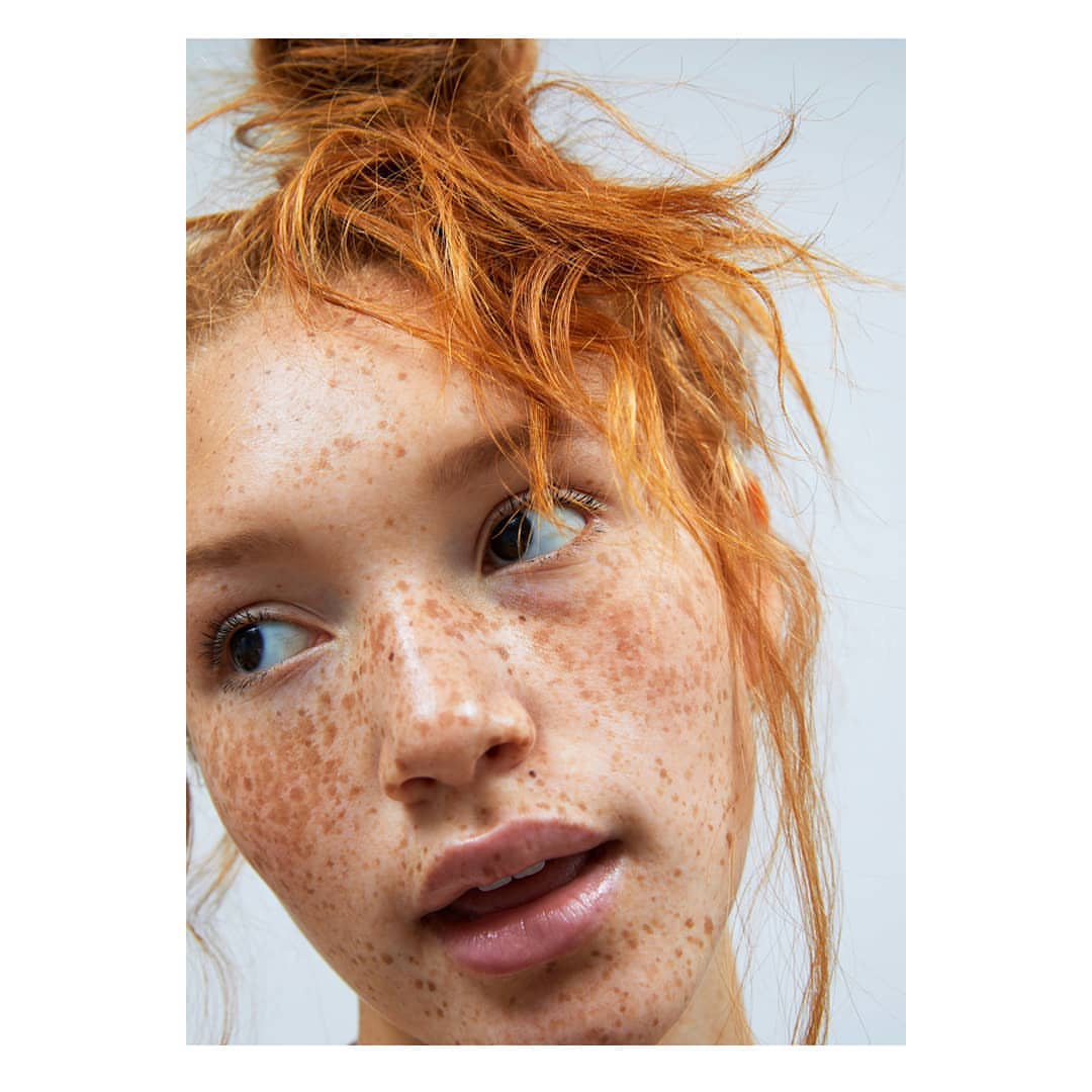 Close-up portrait of a woman with red hair in a messy bun, light skin, and freckles, looking off-camera, set against a clean white backdrop.