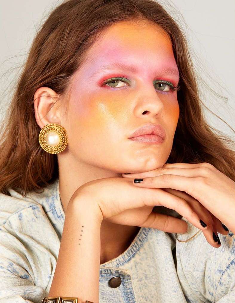 Close-up portrait of a woman with colorful makeup and gold statement earrings, set against a neutral background, highlighting beauty and fashion.