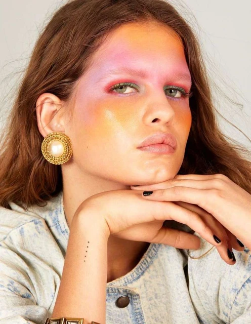 Close-up portrait of a woman with colorful makeup and gold statement earrings, set against a neutral background, highlighting beauty and fashion.