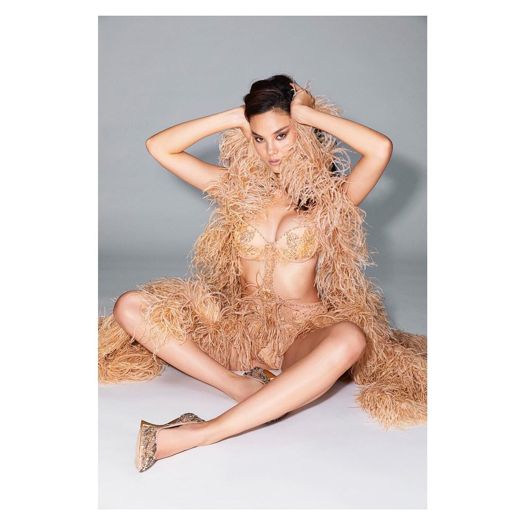 Woman in lingerie and feather wrap poses in studio, seated with hands near her head. Beige tones and soft lighting.