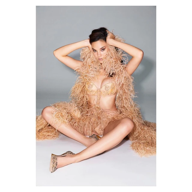 Woman in lingerie and feather wrap poses in studio, seated with hands near her head. Beige tones and soft lighting.