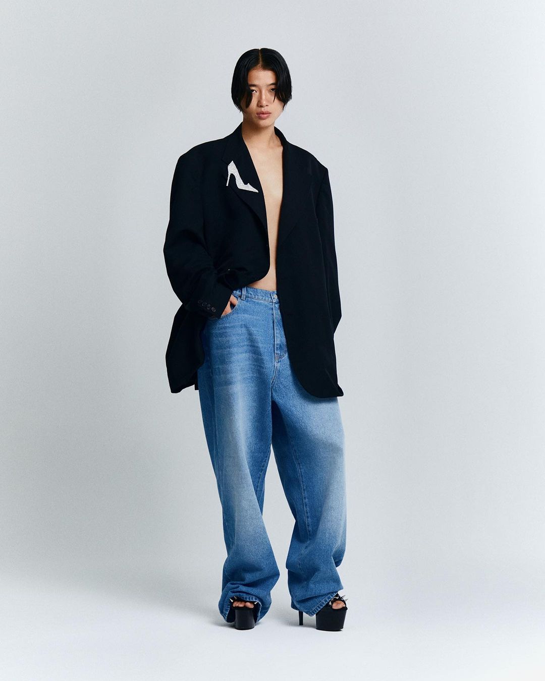 Full-length shot of a person with dark hair wearing oversized blue jeans, a black blazer, and platform heels in a studio setting.