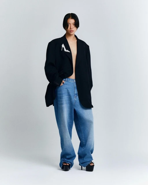 Full-length shot of a person with dark hair wearing oversized blue jeans, a black blazer, and platform heels in a studio setting.