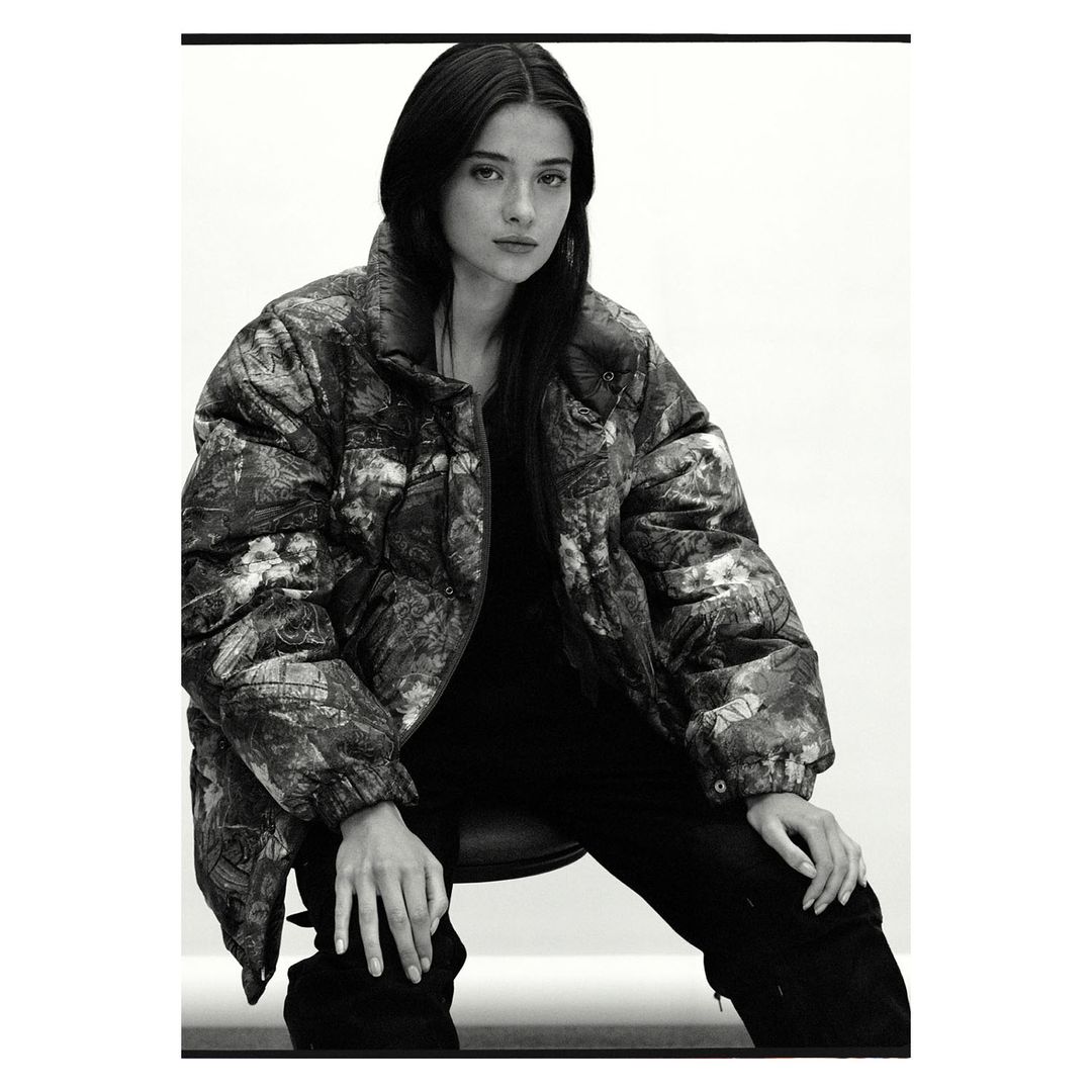 Monochrome portrait of a woman with long, dark hair wearing a patterned puffer jacket, seated against a bright, neutral backdrop.