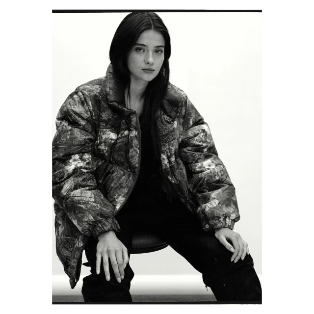 Monochrome portrait of a woman with long, dark hair wearing a patterned puffer jacket, seated against a bright, neutral backdrop.