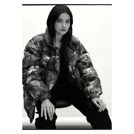Monochrome portrait of a woman with long, dark hair wearing a patterned puffer jacket, seated against a bright, neutral backdrop.