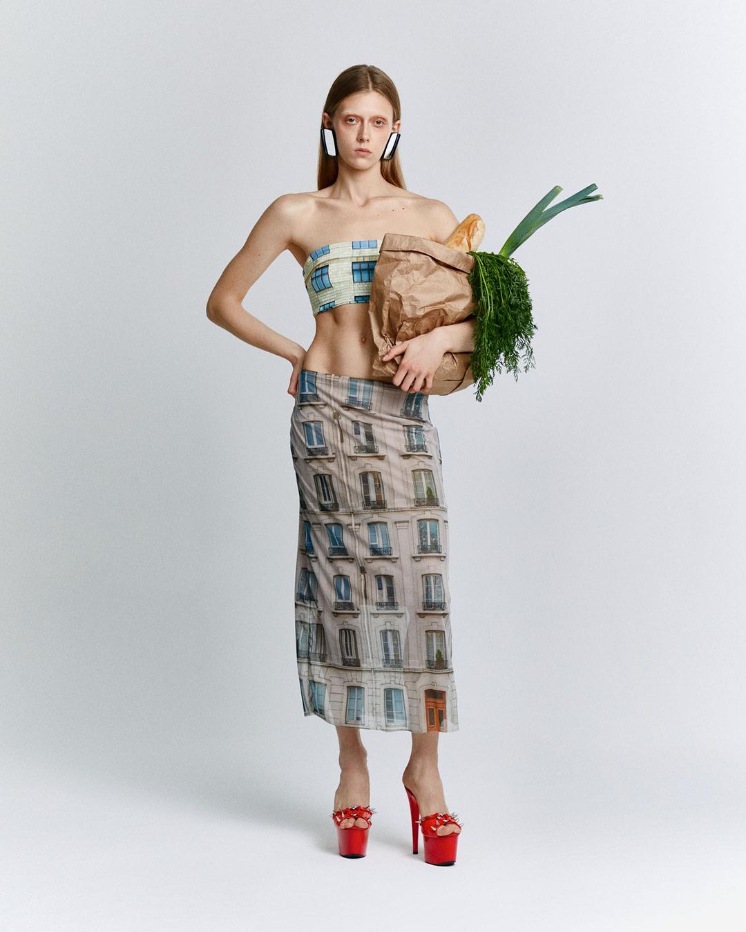 Young woman strikes a pose in fashion shot, modeling a printed top and skirt ensemble with a grocery bag against a white backdrop.