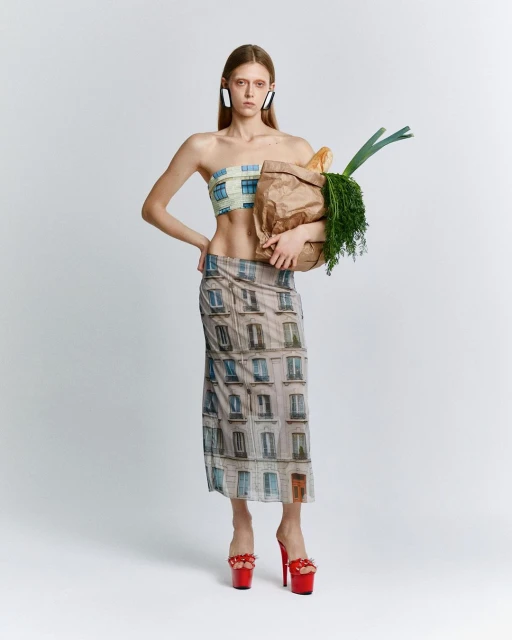 Young woman strikes a pose in fashion shot, modeling a printed top and skirt ensemble with a grocery bag against a white backdrop.