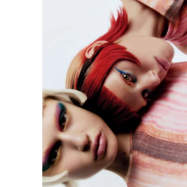 Two adult female faces pressed together, close up portrait, one woman with short red and blonde hair, colorful makeup, and a patterned shirt.