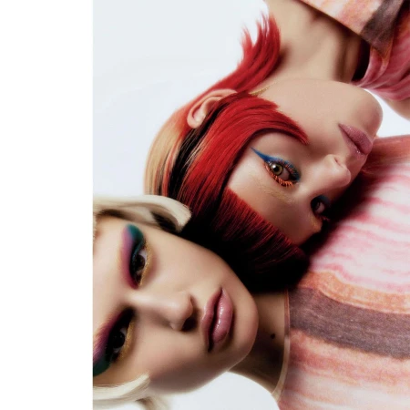 Two adult female faces pressed together, close up portrait, one woman with short red and blonde hair, colorful makeup, and a patterned shirt.