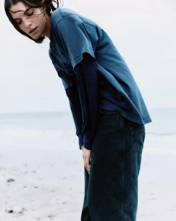 Model in blue t-shirt and dark jeans leaning forward outdoors by the ocean by Carl Diner