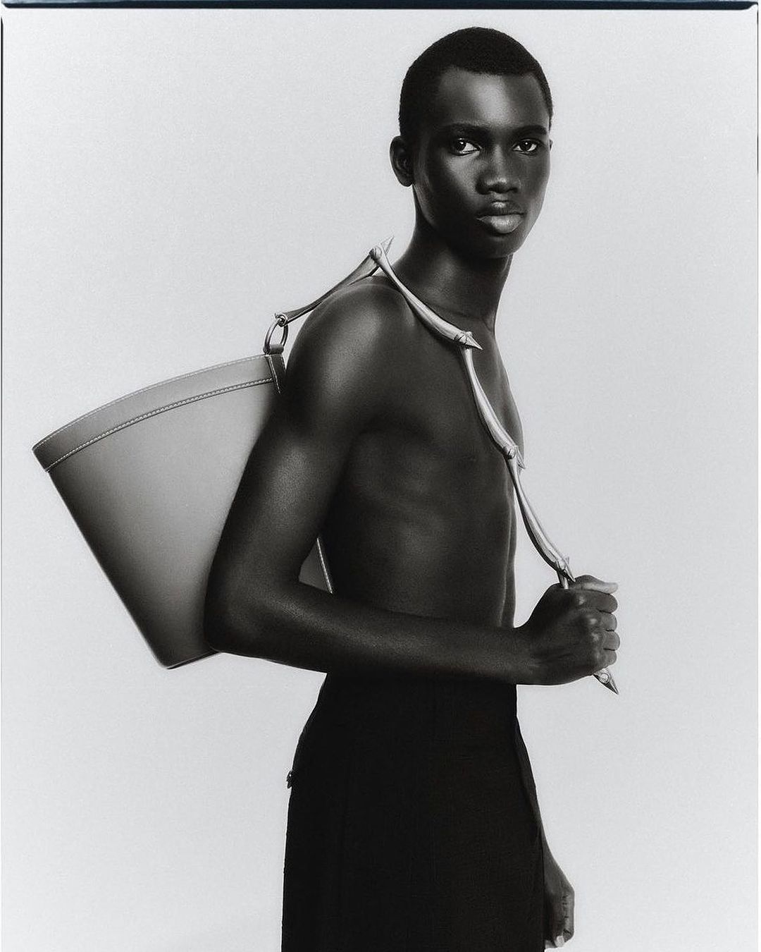 Black male model poses topless with a gray purse, set against a plain white backdrop in a studio fashion shot. Black and white tonality.