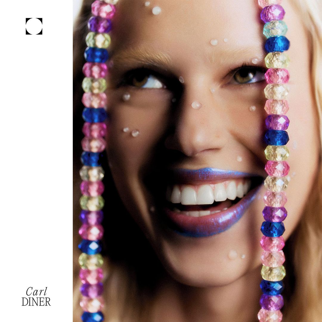 Close-up of a woman with fair skin and blue lipstick smiling with beaded strings, small white decorative dots and a white background.