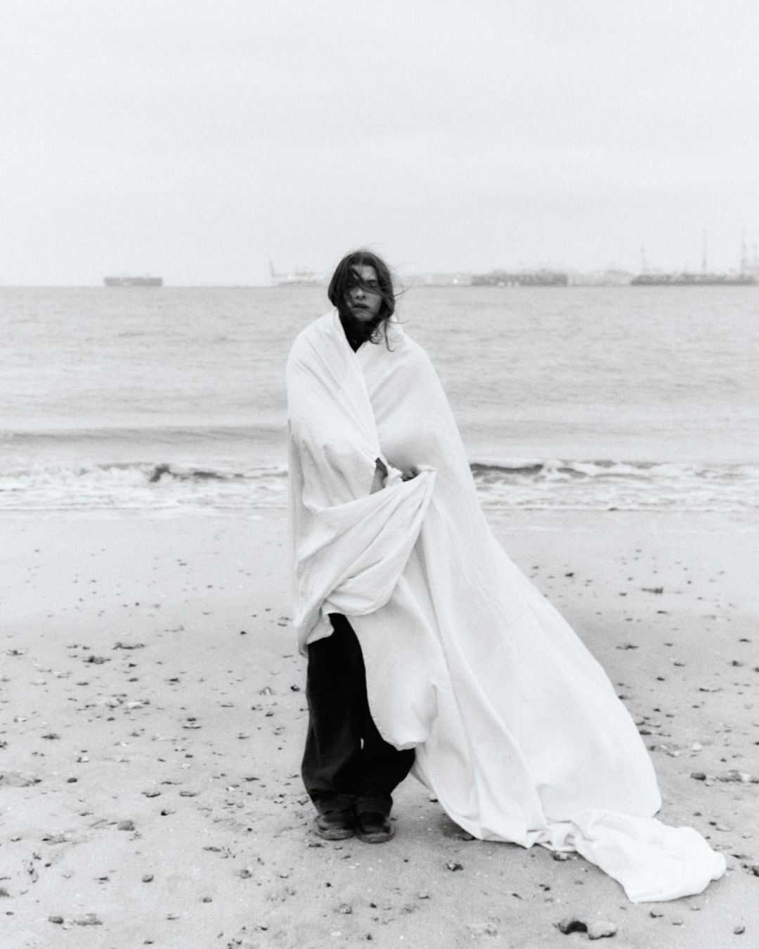 A young adult stands on a sandy beach covered in a white sheet against a cloudy sky, with the ocean and distant ships in the background.