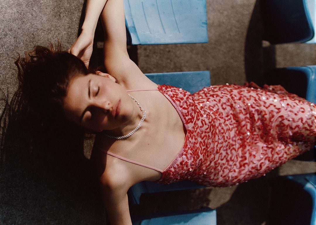 Woman in a red sequin dress lying on blue plastic seats with her eyes closed, photographed from above by Carl Diner in Paris.