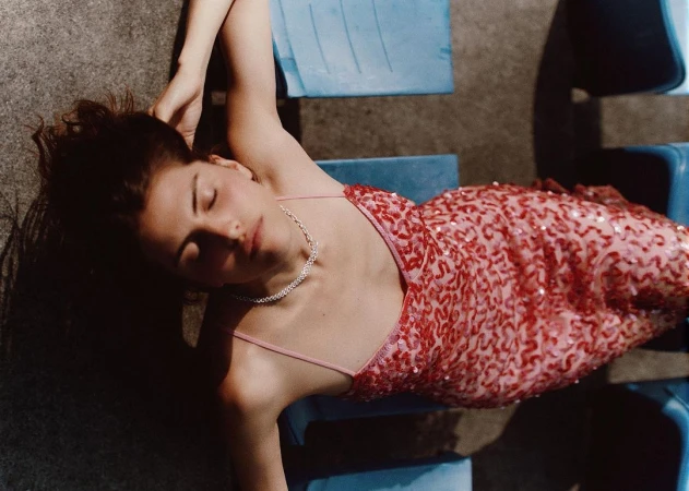Woman in a red sequin dress lying on blue plastic seats with her eyes closed, photographed from above by Carl Diner in Paris.