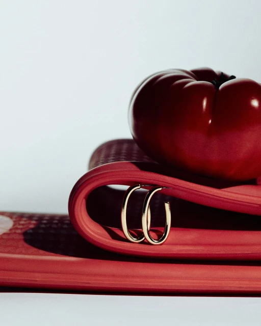 Elegant gold hoop earrings displayed on a vibrant red surface with a glossy red pepper adding contrast, creating a beauty product shot.