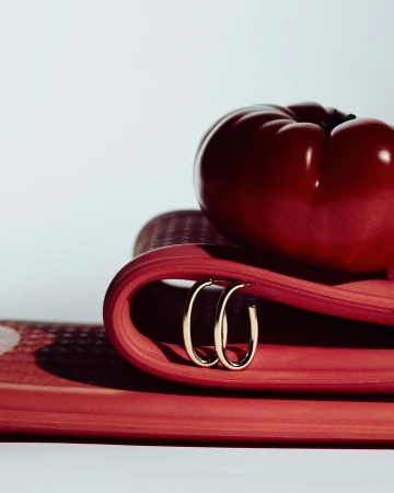 Elegant gold hoop earrings displayed on a vibrant red surface with a glossy red pepper adding contrast, creating a beauty product shot.