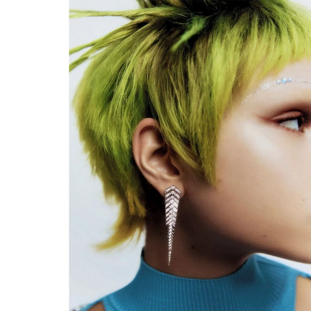 Close-up of a woman with short, bright green hair and a diamond earring. Her skin is light and she is wearing a blue top.
